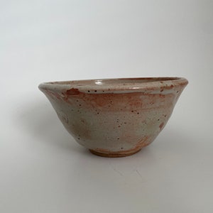 May include: A small, handmade ceramic bowl with a light brown glaze and a speckled finish. The bowl has a rounded shape and a slightly flared rim.