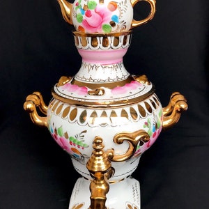 May include: A white porcelain samovar with gold accents and floral designs. The samovar has a teapot on top with a pink base and a white lid with gold trim. The teapot has a floral design with pink, green, and blue accents.
