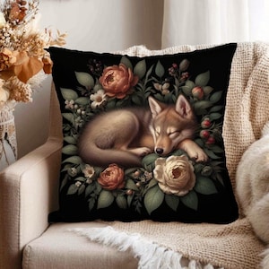 May include: A decorative square pillow with a black background featuring a sleeping wolf cub surrounded by flowers and foliage in shades of brown, peach, and green. The pillow is placed on a beige armchair.