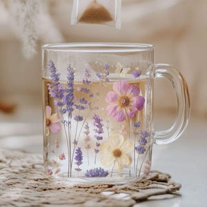 May include: A glass mug with a floral design filled with tea. A tea bag is being lowered into the mug.