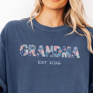 May include: A dark blue short-sleeve t-shirt featuring the word GRANDMA in a floral pattern with white text underneath that reads EST. 2026. The shirt has a crew neckline and a relaxed fit, worn with a light blue denim jacket and denim shorts.