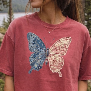 Op de afbeelding: Een baksteenrood T-shirt met korte mouwen en een grote vlinderprint op de borst. De vlinder is verdeeld in twee verschillende patronen: de linkervleugel heeft een blauw-wit paisley-ontwerp, terwijl de rechtervleugel een rood-wit bloemenpatroon toont.