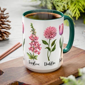 May include: White ceramic mug with a green handle and rim, filled with a dark liquid. The mug is decorated with watercolor illustrations of pink and purple flowers, including foxglove, dahlia, and tulip. The names of the flowers are written below each illustration.