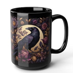 May include: Black ceramic mug featuring a detailed illustration of a raven perched within a floral wreath. The raven is depicted in shades of purple and black, set against a golden background. The flowers are in shades of purple, gold, and brown.