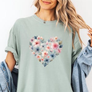 May include: A pale green t-shirt with a heart-shaped floral design. The heart is made up of pink, blue, and white flowers with green leaves. The shirt has short sleeves and a loose fit.