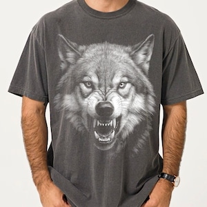 May include: A charcoal gray short-sleeve t-shirt featuring a large, detailed graphic print of a snarling wolf face on the front. The wolf is depicted in shades of gray and white with bared teeth and focused eyes, centered on the chest of the shirt.