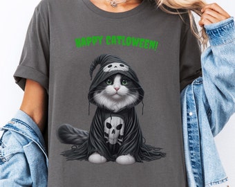 Puede incluir: Camiseta gris oscuro con un gato de dibujos animados vestido con una capa con capucha negra con detalles de calaveras. El gato tiene pelaje blanco, ojos verdes y una cara en blanco y negro. El texto verde dice "HAPPY CATOWEEN!"