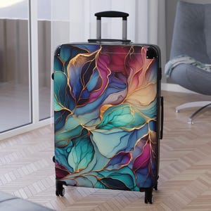 Puede incluir: Una maleta con ruedas negra con un diseño floral abstracto en tonos turquesa, morado y dorado. El diseño se asemeja a hojas. La maleta tiene un asa retráctil y cuatro ruedas.