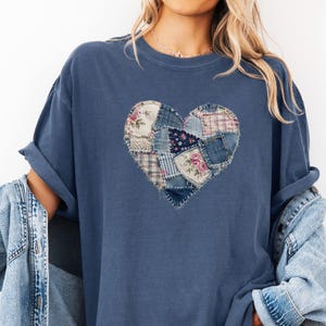 May include: A slate blue short-sleeve t-shirt featuring a central heart graphic composed of various patchwork fabric patterns, including floral prints, plaid, and denim textures, layered over a light blue denim button-down shirt.