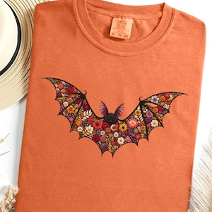 May include: An orange t-shirt with a floral bat design. The bat is outlined in black and filled with colorful flowers. A straw hat and dried plants are in the background. The shirt is a casual, comfortable style.
