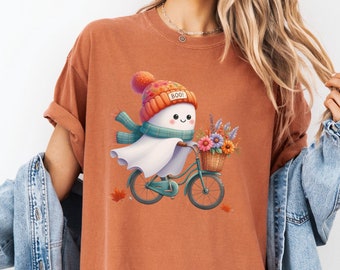 May include: Rust-colored t-shirt featuring a whimsical illustration of a ghost riding a teal bicycle. The ghost wears a knit hat with "BOO" on it and a scarf, carrying flowers in a basket. Autumn leaves are scattered around the bicycle.