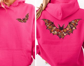 May include: A bright pink hooded sweatshirt featuring a small bat graphic with floral wings on the left chest and a large, matching bat graphic with intricate floral patterns on the back. The hoodie includes a front kangaroo pocket and a hood.