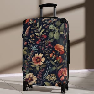 Puede incluir: Una maleta con ruedas con un fondo azul marino oscuro y un estampado floral. El diseño presenta varias flores en tonos naranja, rojo y crema, junto con follaje verde. La maleta tiene un asa y ruedas negras.
