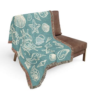 May include: A teal throw blanket with a repeating pattern of white seashells and starfish. The blanket has a fringed edge with brown and orange threads. The blanket is draped over a brown sofa.