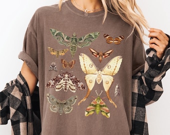 May include: A brown t-shirt with a vintage illustration of various moths and butterflies. The design features a variety of colors and patterns, including green, brown, yellow, and white.