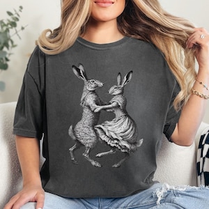 Puede incluir: Una camiseta gris antracita con un estampado gráfico de dos liebres bailando. Las liebres están dibujadas en blanco y negro y llevan ropa.