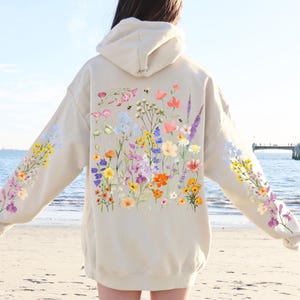 May include: A cream-coloured hoodie with a floral print featuring a variety of colourful flowers, including pink, purple, yellow, and blue. The hoodie has a drawstring hood and a kangaroo pocket.