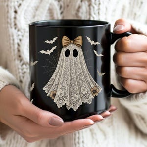May include: Black ceramic mug featuring a ghost design with a lace-like appearance, a bow, and floral accents. The design includes bats and a spiderweb. The mug is held by a person wearing a white sweater.