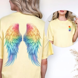Puede incluir: Una camiseta amarilla clara con un par de alas de arcoíris coloridas en la espalda. Una imagen más pequeña muestra una camiseta a juego con un pequeño diseño de arcoíris en la parte delantera. Las alas son un diseño de acuarela con tonos de rojo, naranja, amarillo, verde, azul y morado.