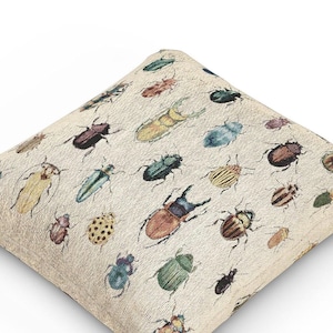 May include: A beige cushion with a colourful pattern of various beetles and bugs. The cushion is made of a soft, textured fabric and features a variety of insect designs.