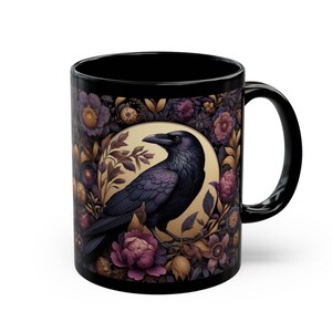 May include: Black ceramic mug featuring a detailed illustration of a raven perched within a floral design. The raven is depicted in shades of purple and black, surrounded by purple and gold flowers. The mug has a black handle and interior.