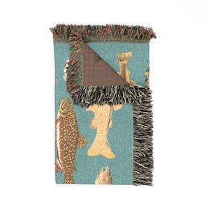 May include: A folded turquoise throw blanket featuring a repeating pattern of various fish illustrations in shades of brown, yellow, and red. The blanket has a fringed edge with a mix of colors. The underside of the blanket is brown.