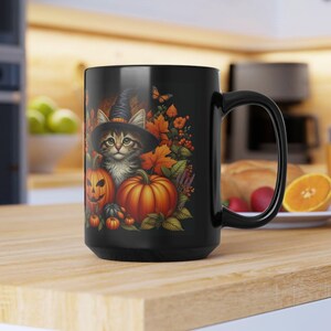 May include: Black ceramic mug featuring a Halloween-themed design. The design includes a kitten wearing a witch hat, pumpkins, and autumn leaves. The mug has a large handle and is sitting on a wooden surface.