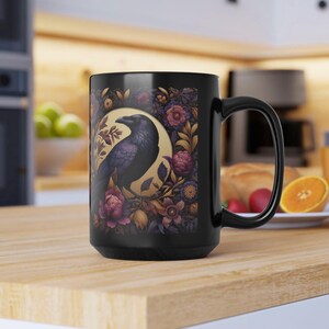 May include: Black ceramic mug featuring a detailed illustration of a raven within a golden circle, surrounded by floral elements in shades of purple, pink, and gold. The mug has a large handle and is suitable for hot or cold beverages.
