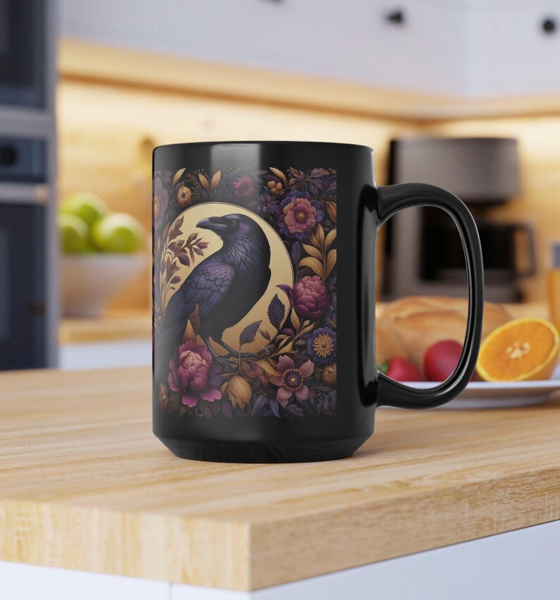 May include: Black ceramic mug featuring a detailed illustration of a raven within a golden circle, surrounded by floral elements in shades of purple, pink, and gold. The mug has a large handle and is suitable for hot or cold beverages.