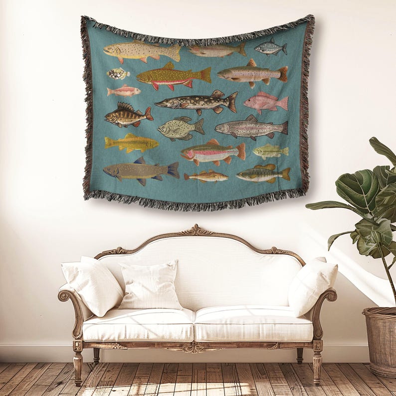 May include: A teal throw blanket featuring illustrations of various fish species. The blanket has a fringed edge and is displayed on a white wall above a white sofa. The fish are depicted in shades of yellow, red, and gray.
