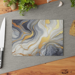 May include: Rectangular cutting board with a marble design. The board features a swirling pattern of gray, white, and gold. The gold veins create a luxurious look. The board is placed on a gray surface with a knife on the left side.