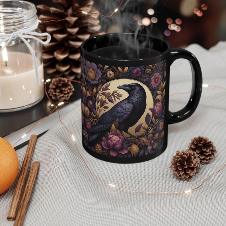 May include: Black ceramic mug featuring a detailed illustration of a raven surrounded by purple and gold flowers. The raven is centered on a gold background. The mug has a black handle and is filled with a steaming dark liquid.