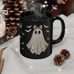 May include: Black ceramic mug featuring a ghost design with a lace-like shroud and a bow. The ghost has two oval eyes and is surrounded by bats and spiderwebs. The mug is a Halloween-themed product.