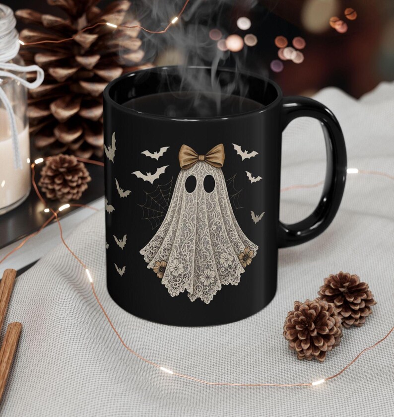 May include: Black ceramic mug featuring a ghost design with a lace-like shroud and a bow. The ghost has two oval eyes and is surrounded by bats and spiderwebs. The mug is a Halloween-themed product.