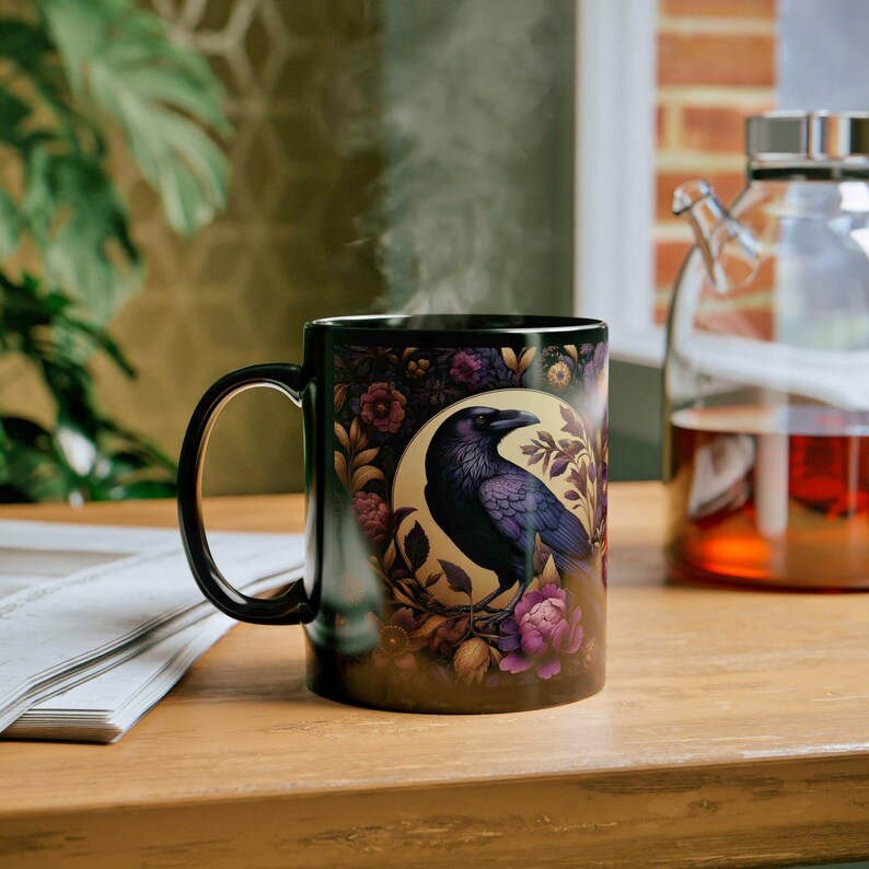 May include: Black ceramic mug featuring a detailed illustration of a raven perched on a branch, surrounded by floral elements in shades of purple, gold, and brown. The mug has a black handle and a glossy finish, perfect for hot beverages.