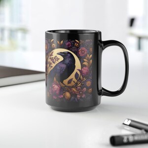 May include: Black ceramic mug featuring a detailed illustration of a raven perched on a branch, set against a gold circle and surrounded by floral elements in shades of purple, pink, and gold. The mug has a large handle.