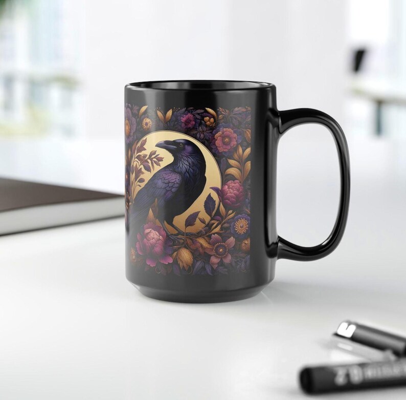 May include: Black ceramic mug featuring a detailed illustration of a raven perched on a branch, set against a gold circle and surrounded by floral elements in shades of purple, pink, and gold. The mug has a large handle.