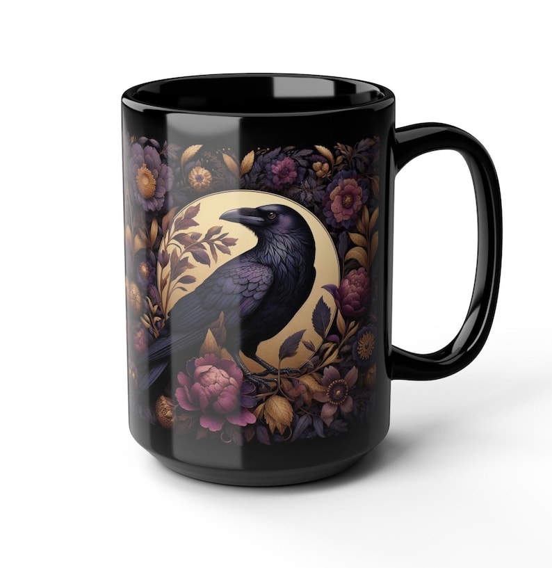 May include: Black ceramic mug featuring a detailed illustration of a raven perched within a floral wreath. The raven is depicted in shades of purple and black, set against a golden background. The flowers are in shades of purple, gold, and brown.