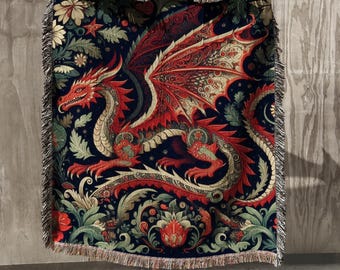 Puede incluir: Una manta tejida con un diseño de dragón rojo y alas color crema, sobre un fondo azul oscuro con detalles florales y de hojas. La manta tiene un borde con flecos y está hecha de un material suave.