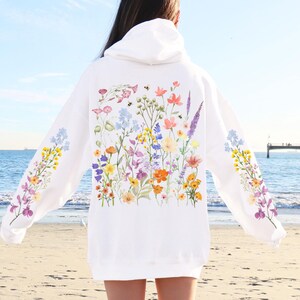 May include: White hoodie with a floral print featuring a variety of colorful flowers and bees. The hoodie has a drawstring hood and a kangaroo pocket.