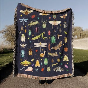 May include: A navy blue throw blanket featuring a colorful pattern of various insects, including butterflies, beetles, and dragonflies. The blanket has a fringed edge and is made of a soft material.