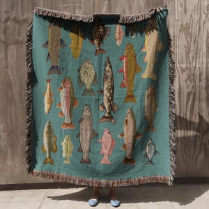 May include: Teal throw blanket featuring various fish illustrations in shades of brown, yellow, pink, and gray. The blanket has a fringed edge and is displayed against a neutral background.