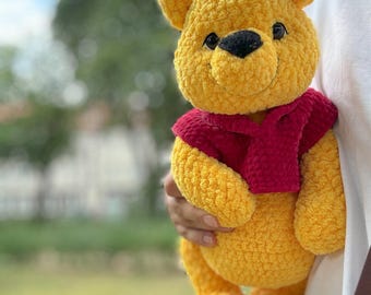 Op de afbeelding: Een gehaakt Winnie de Poeh knuffel met een rode sjaal, vastgehouden in de hand van een persoon.