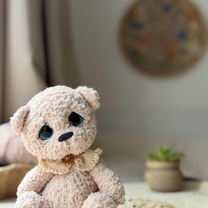 Könnte beinhalten: Ein beigefarbener Plüschteddybär mit einer strukturierten, flauschigen Oberfläche. Der Bär hat große, dunkle Augen mit blauen Akzenten, eine kleine schwarze gestickte Nase und einen hellbeigen gehäkelten Kragen mit einem kleinen braunen Holzknopf.