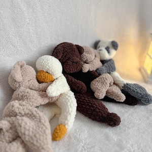 Könnte beinhalten: Eine Sammlung handgefertigter, plüschiger Stofftiere in verschiedenen Farben. Die Spielzeuge umfassen einen hellrosa Bären, eine weiße Ente mit gelben Akzenten, einen braunen Bären, einen beigefarbenen Bären und einen Panda mit grauen und weißen Details. Die Spielzeuge sind auf einer weißen, strukturierten Oberfläche angeordnet.