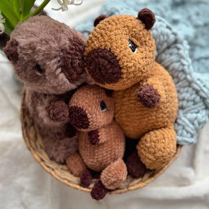 May include: Three crocheted stuffed animals in a wicker basket. The animals are brown and tan with brown eyes and noses. The animals are sitting in a wicker basket on a white surface.