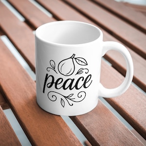 May include: White ceramic mug with a black illustration of a fruit and the word "peace" in a cursive font. The mug has a curved handle and is set against a wooden surface.