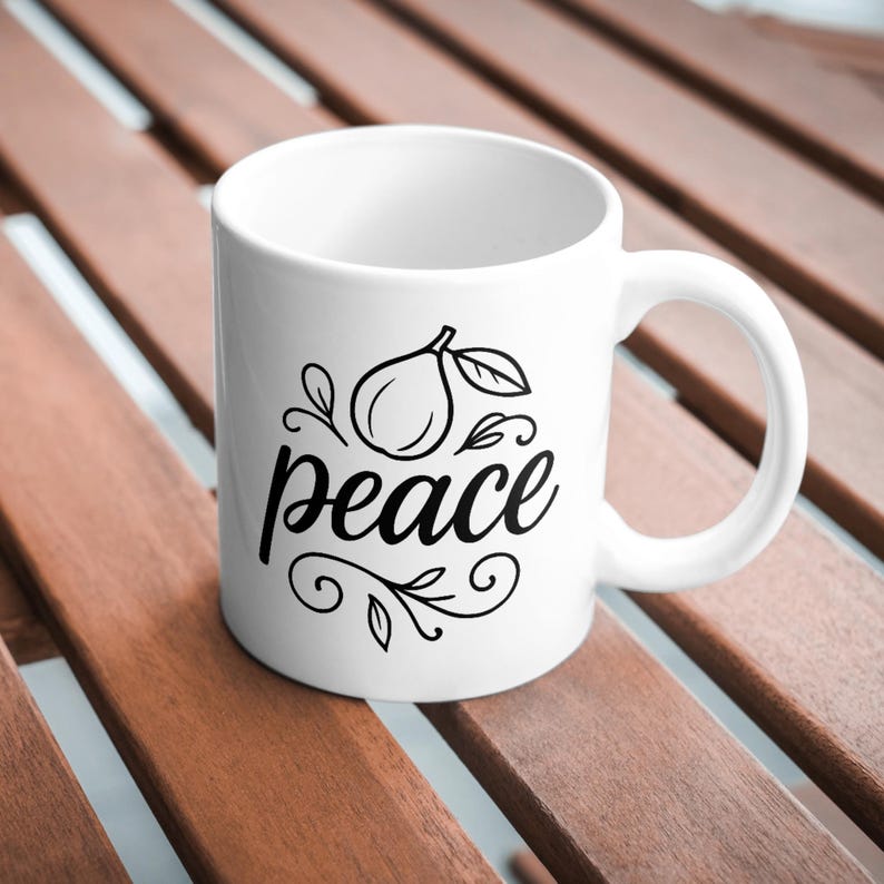 May include: White ceramic mug with a black illustration of a fruit and the word "peace" in a cursive font. The mug has a curved handle and is set against a wooden surface.