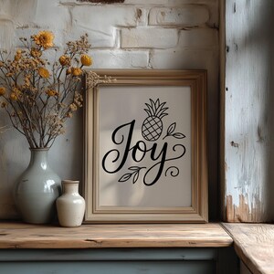 May include: A framed print with the word "Joy" in elegant script, accompanied by a pineapple illustration. The artwork is displayed on a wooden surface, next to a vase with dried flowers and a smaller vase. The background features a brick wall.