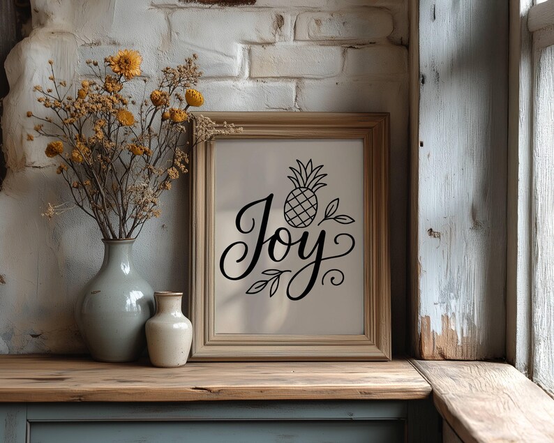 May include: A framed print with the word "Joy" in elegant script, accompanied by a pineapple illustration. The artwork is displayed on a wooden surface, next to a vase with dried flowers and a smaller vase. The background features a brick wall.